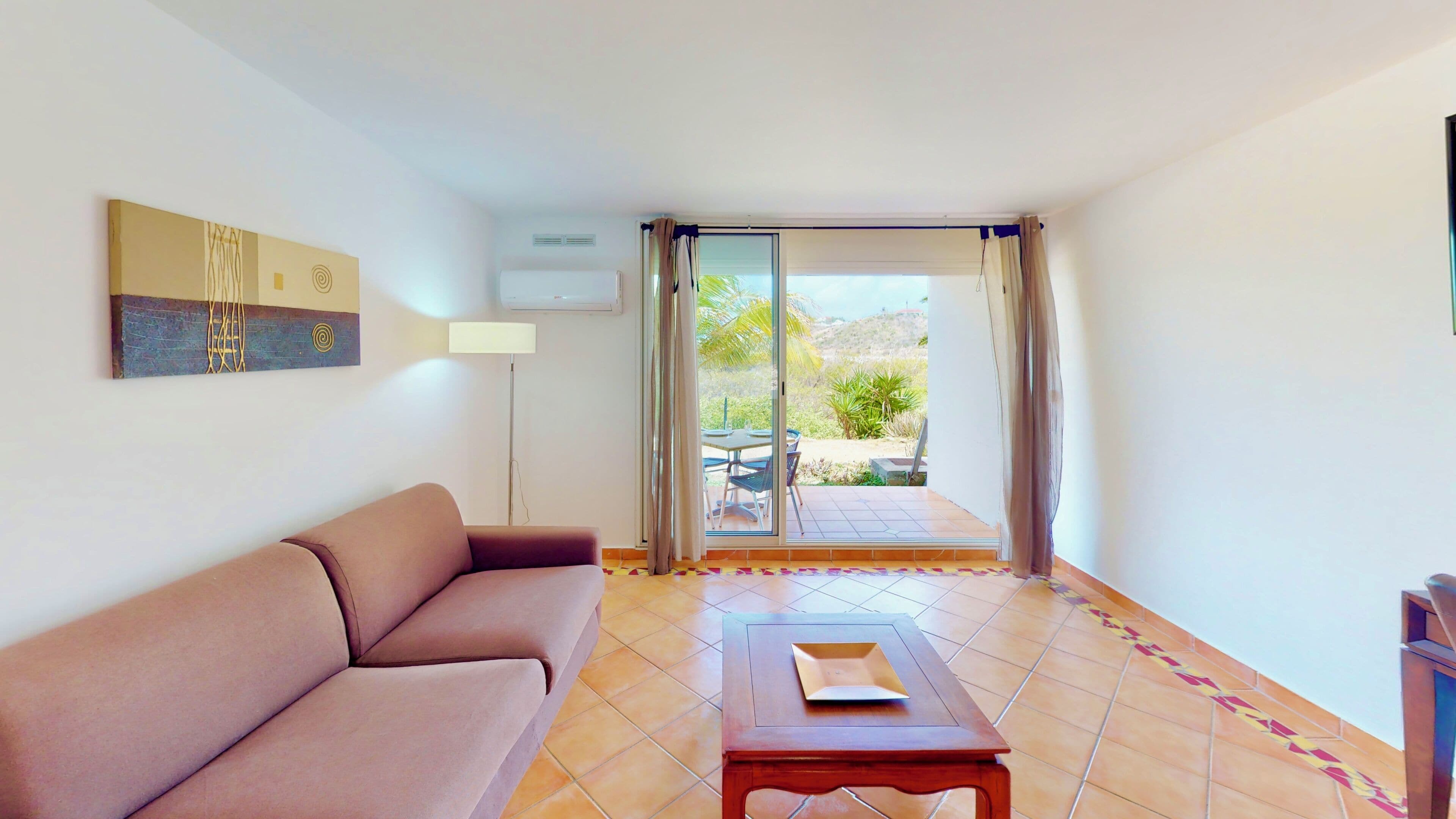 A room with a sofa, a table, and a view through a sliding glass door to an outdoor area with a table and chairs.