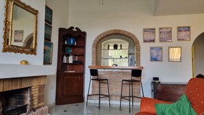 Ein gemütliches Wohnzimmer mit Kamin, einer Ziegelwand, einem hölzernen Bücherregal, zwei schwarzen Sesseln und einem Sofa mit grüner Decke.