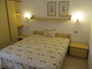 Un dormitorio con una cama, dos mesitas de noche, una mesilla auxiliar, un estante con fotografías enmarcadas y luces fijadas a la pared.
