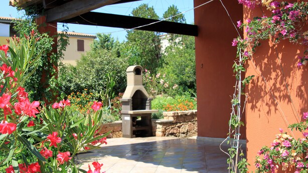 Un patio con una parrilla de piedra, rodeado de plantas en flor y un muro de piedra.