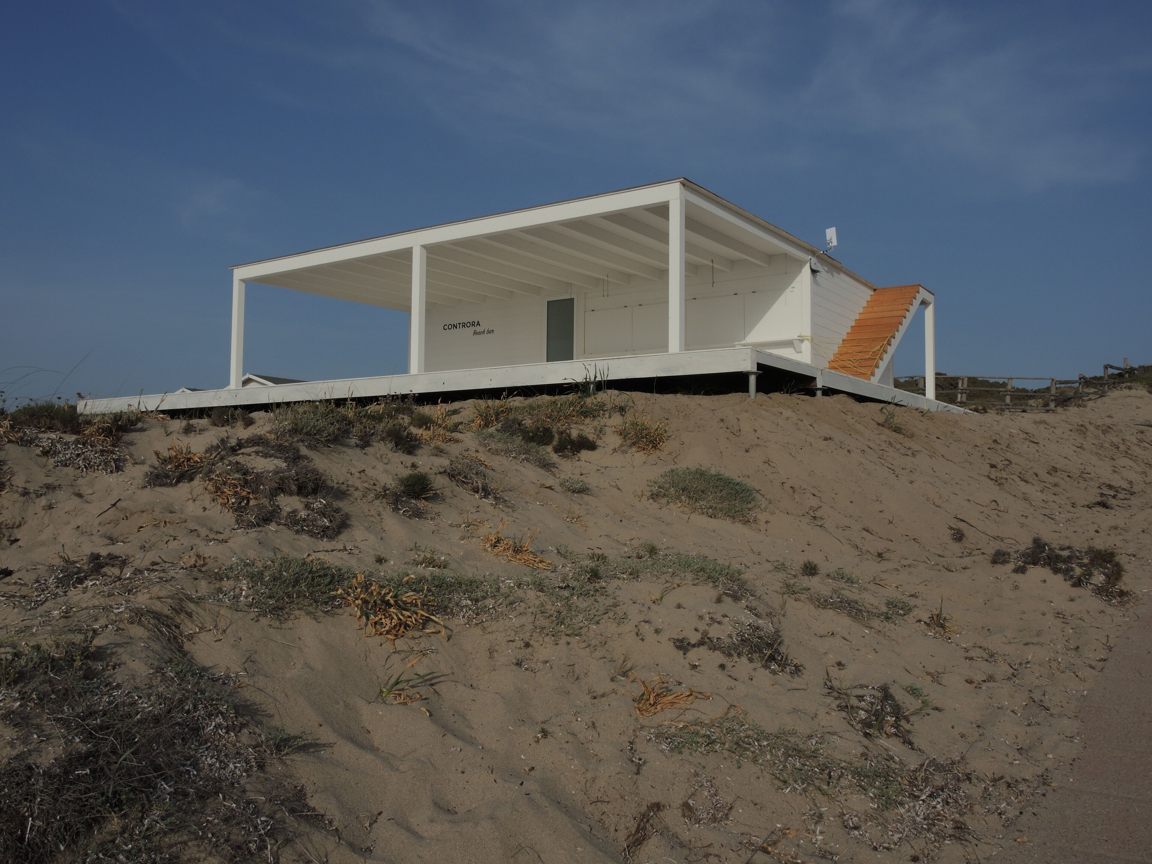A modern beach house with a white exterior, a covered porch, and a wooden staircase leading to the entrance.