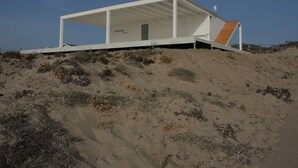 Una casa moderna frente al mar, con fachada blanca, un porche cubierto y una escalera de madera que conduce a la entrada.