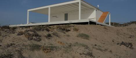 A modern beach house with a white exterior, a covered porch, and a wooden staircase leading to the entrance.