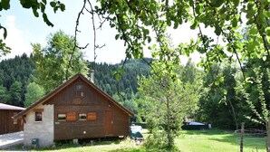 Una cabaña de madera con techo inclinado, un pequeño balcón y una puerta. Hay un patio con césped en frente de la cabaña, y un árbol a la derecha.