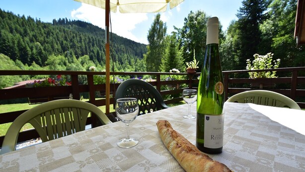 Una mesa con una botella de vino, una copa y una baguette sobre un mantel a cuadros, ubicada al aire libre con vistas a una montaña boscosa.