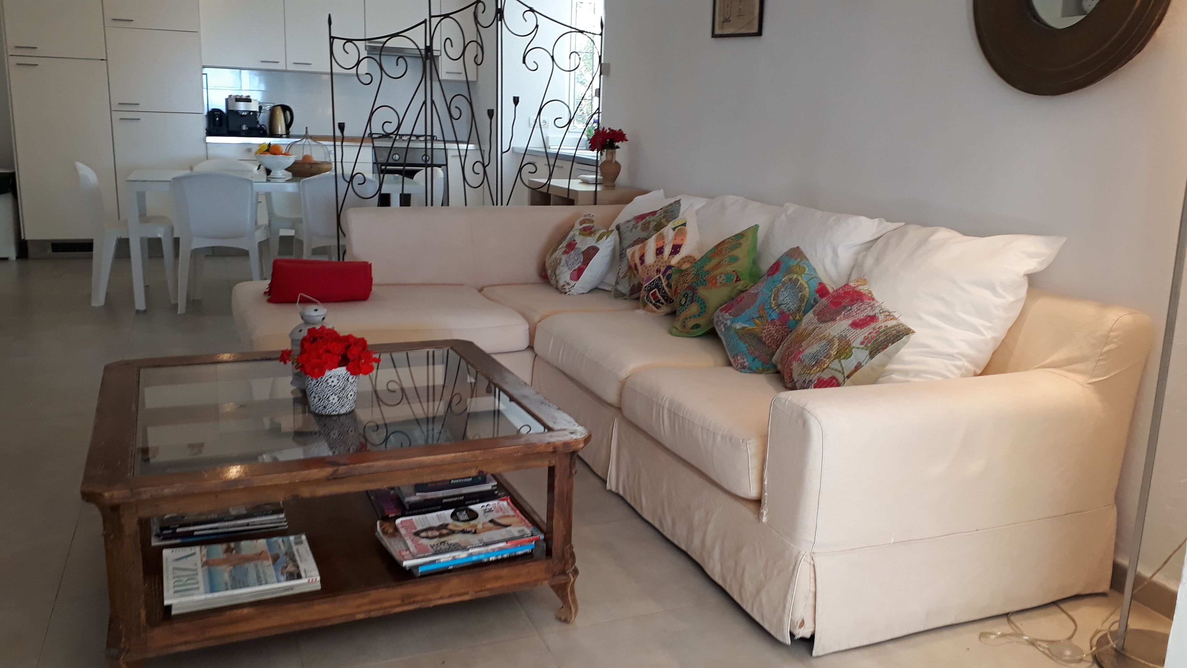 A living room with a white sofa, colorful cushions, a glass coffee table with magazines, and a red cushion on the sofa.