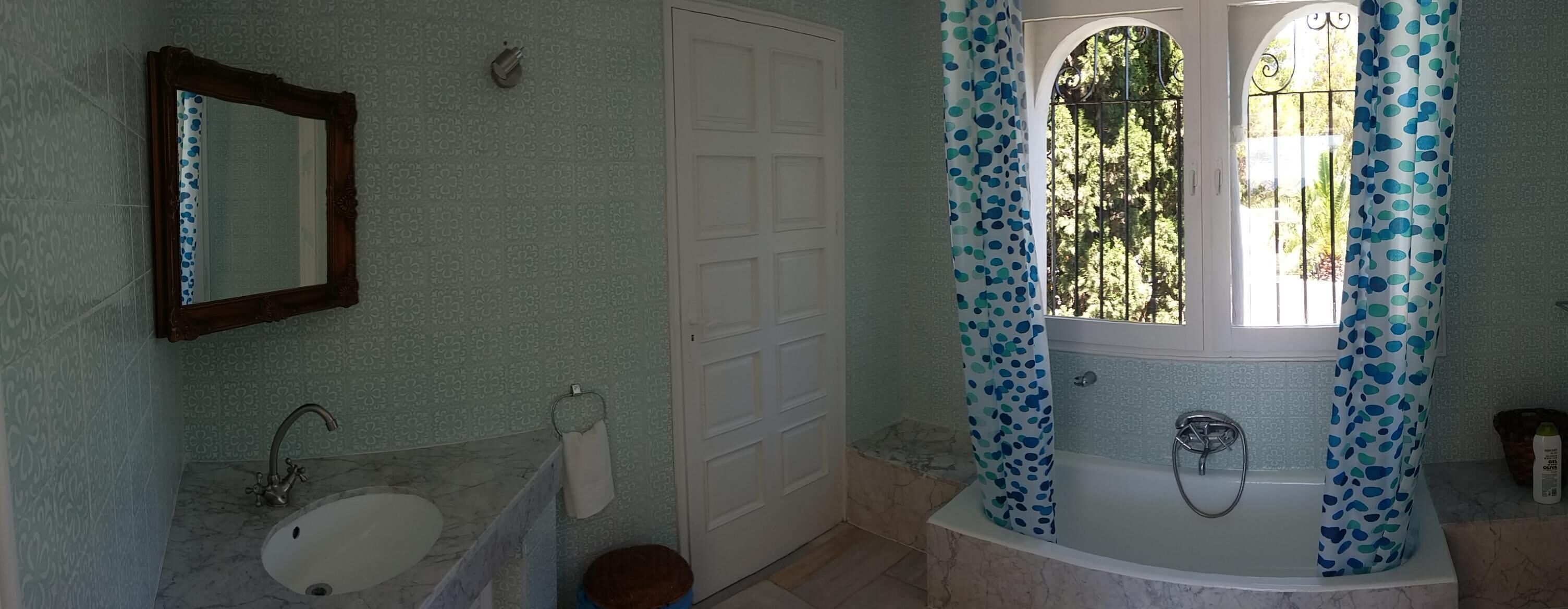 A bathroom with a marble sink, a white door, a bathtub with blue polka dot curtains, and a window with a view of trees.