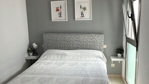 Ett sovrum med en säng, två inramade konstverk, ett nattduksbord, ett fönster med persienner och en liten hylla med en växt.