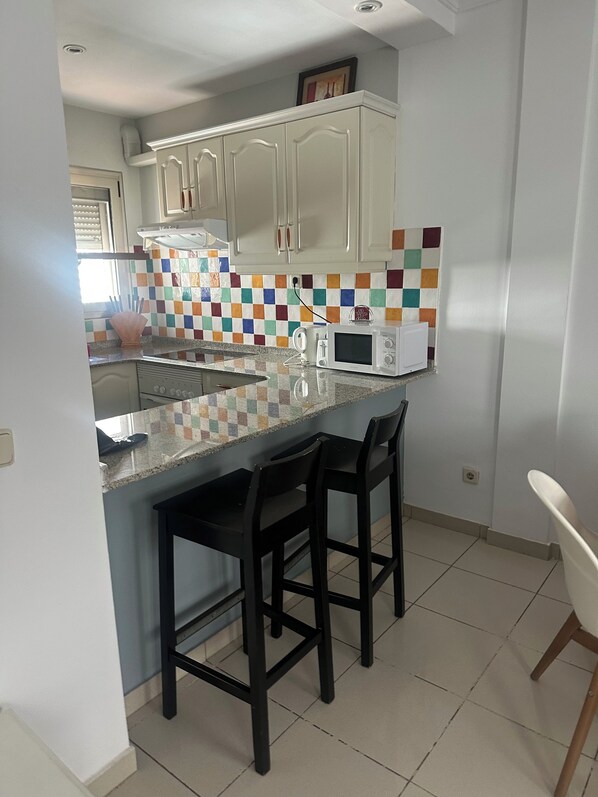 A kitchen with white cabinets, a colorful checkered backsplash, a microwave, and a bar area with two stools.