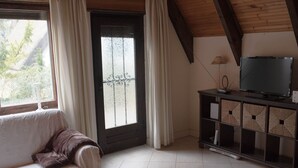 Une pièce avec un canapé blanc, un téléviseur posé sur un meuble en bois, une porte en verre dépoli et une fenêtre avec des rideaux.
