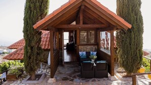 A wooden gazebo with a stone floor, a seating area with cushions, and tall trees on either side.