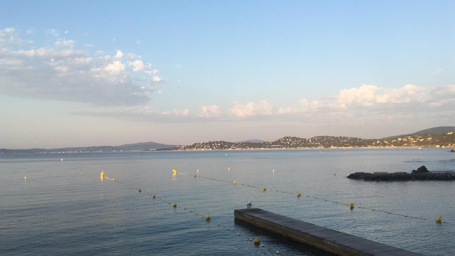 A coastal view with calm waters, a concrete pier, and distant hills with buildings.