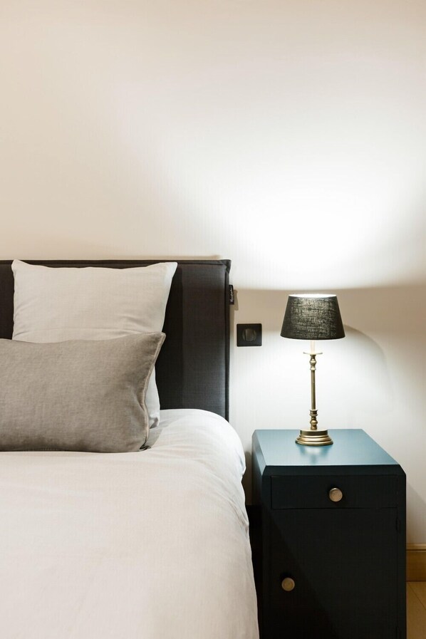 A neatly made bed with a white comforter and two pillows, a small bedside table with a lamp, and a dark-colored nightstand.