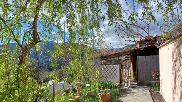 A stone pathway leading to a wooden building with a sloped roof, surrounded by lush greenery and a mountainous backdrop.