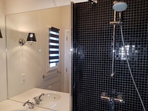 Een badkamer met een witte wasbak, een spiegel, een douchegedeelte met zwarte tegels en een raam met zonwering.