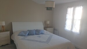 Un dormitorio con una cama, dos mesitas de noche, una lámpara colgante y un ventanal con cortinas.