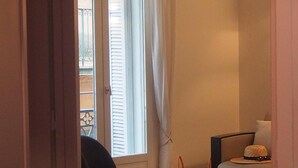 A room with a wooden floor, a sofa, a chair, a small table, and a window with white curtains. - Appart Aix Mazarin 3 stars-T2 with air conditioning in the historic city centre (Aix-en-Provence)