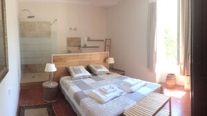Quarto com cabeceira de madeira, duas luminárias de cabeceira, cama com colcha xadrez, mesa de cabeceira pequena com abajur, espelho, área de chuveiro e janela com cortinas.