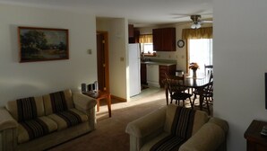 Una sala de estar con un sofá y sillones, una mesa de comedor con sillas, una zona de cocina con armarios y electrodomésticos, y un cuadro en la pared.