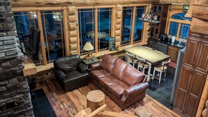 A log cabin interior with a stone fireplace, leather sofa, dining table, and kitchen area.