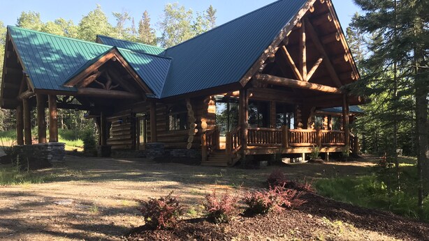 A log cabin with a porch and a covered deck, surrounded by a forested area.