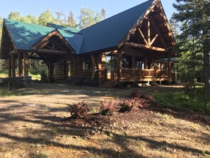 A log cabin with a porch and a covered deck, surrounded by a forested area.