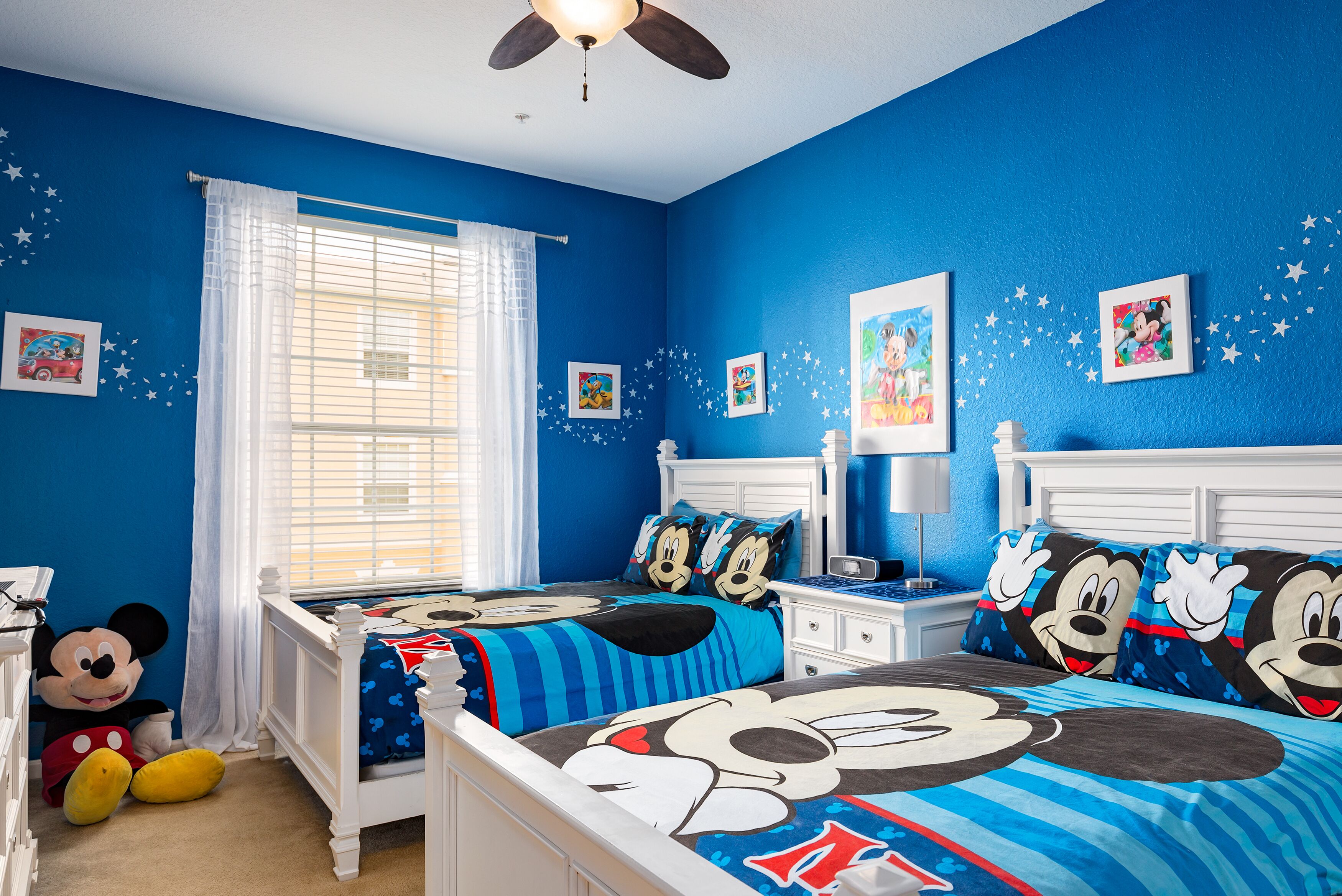 Une chambre à coucher aux couleurs bleues, avec du linge de lit à l’effigie de Mickey Mouse, un grand Mickey en peluche et des décorations en forme d’étoiles.