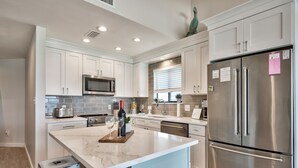 A modern kitchen with white cabinets, a stainless steel refrigerator, a marble island, and a built-in microwave.