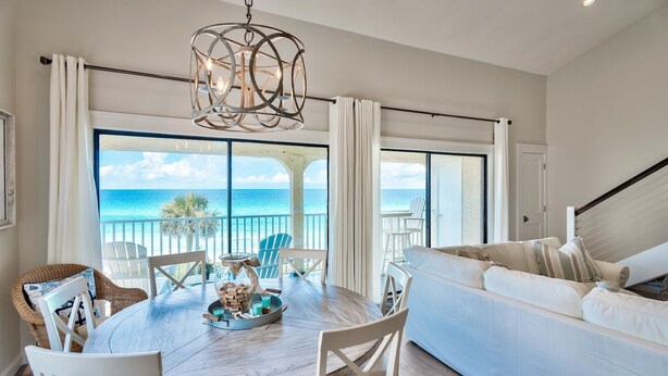 A spacious living area with a large round table, white chairs, and a sofa. A balcony with a view of the ocean is visible through large glass doors.