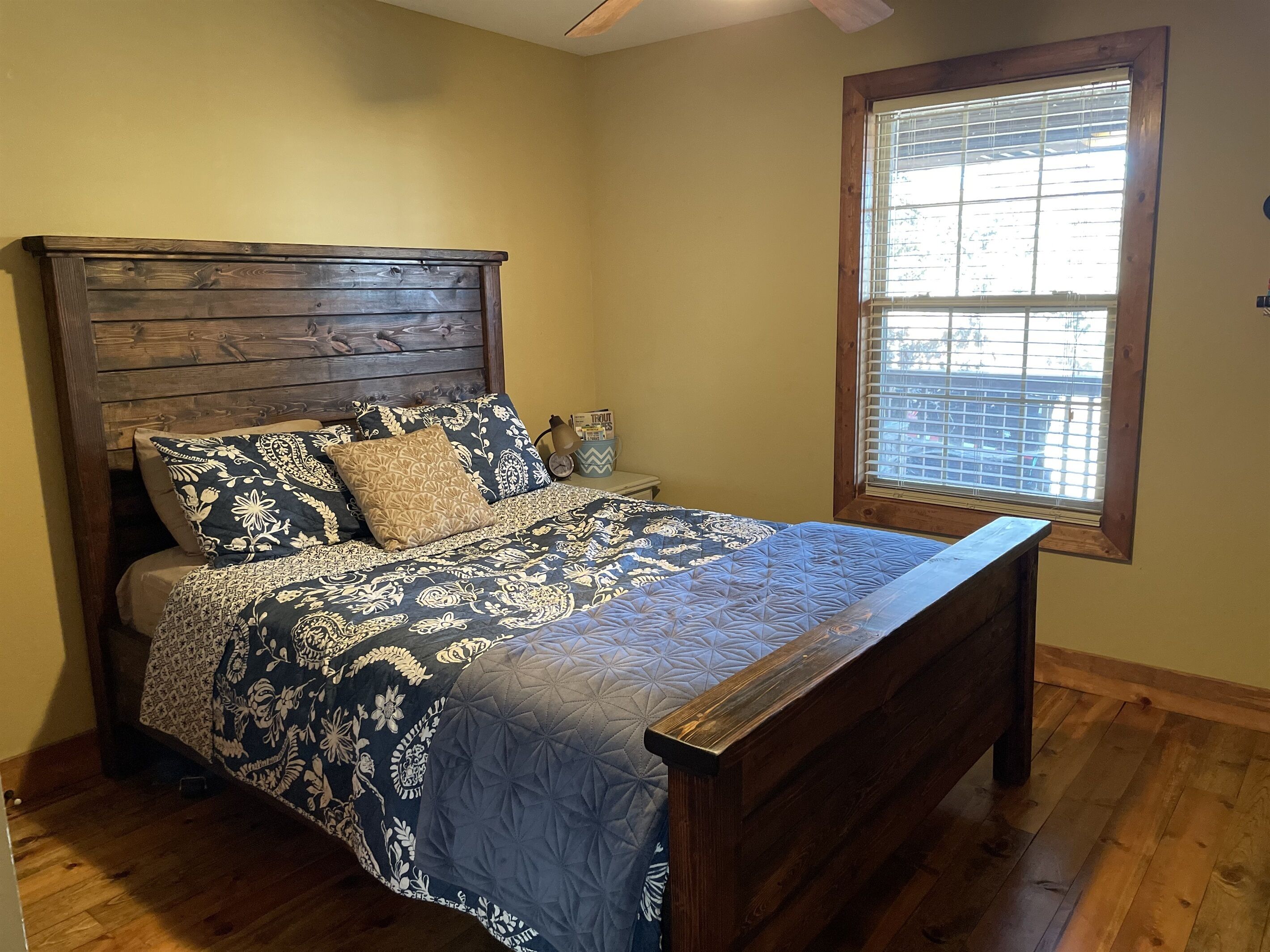Un dormitorio con cabecera de madera, ropa de cama estampada, una mesita de noche con lámpara y una ventana con persianas.