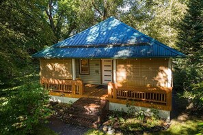 A small, single-story cabin with a blue metal roof, a front porch with wooden railings, and a brick fire pit in front. - Clean, Private, Historic, Downtown Bozeman Home (Bozeman)