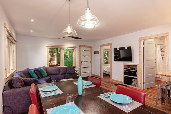 A modern living room with a dark wooden dining table set for four, a sofa with colorful cushions, a ceiling fan, and a TV mounted on the wall. - Clean, Private, Historic, Downtown Bozeman Home (Bozeman)