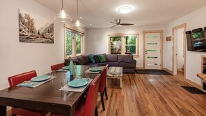 Uma sala de jantar com mesa de madeira, cadeiras vermelhas e um sofá. Há janelas com vista para o verde lá fora.