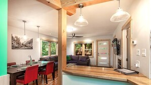 Cozinha moderna com vigas de madeira, área de refeições com cadeiras vermelhas e vista para uma sala de estar com sofá e televisão.