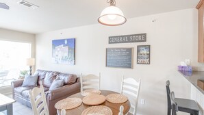 Comedor con una mesa redonda, sillas blancas y individuales tejidos. Hay un sofá de cuero marrón y un letrero en la pared que dice "GENERAL STORE".