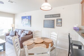 A dining area with a round table, white chairs, and woven placemats. There is a brown leather couch and a wall-mounted sign that reads "GENERAL STORE."