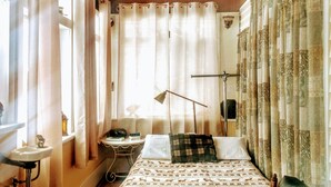A bedroom with a wooden ceiling, a bed with patterned bedding, a black leather chair, a bedside table with a lamp, and a window with sheer curtains.