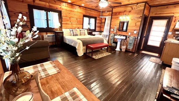 A wooden cabin interior with a bed, bedside table, lamp, and a vase with flowers.