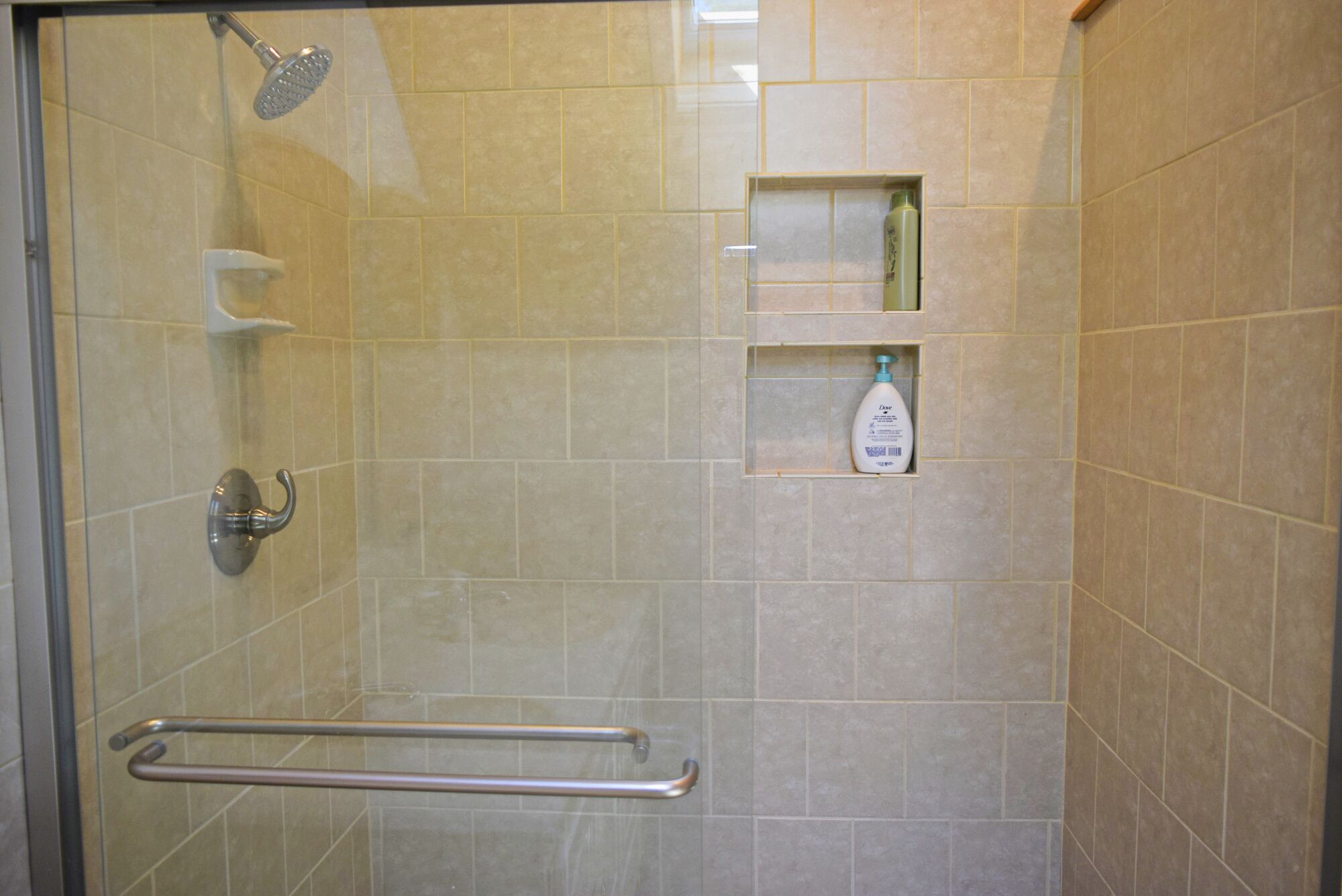 A glass-enclosed shower with a handheld showerhead, a grab bar, and a shelf with toiletries.