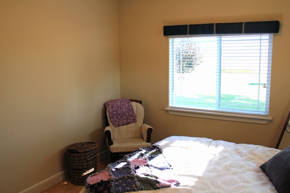 A bedroom with a bed, a chair with a blanket, a window with blinds, and a trash bin. - Close To Everything! Well Equipped, Centrally Located House Near Coeur D'alene. (Post Falls)