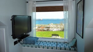 Ein Zimmer mit an der Wand befestigtem Fernseher, einem Fenster mit Blick auf das Meer und Gebäude sowie einer Bank mit gemusterten Kissen.