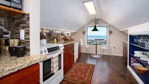 Eine Küche mit Herd, Backofen und verschiedenen Geräten. Ein Esstisch mit Stühlen und Blick auf das Meer durch ein Fenster.