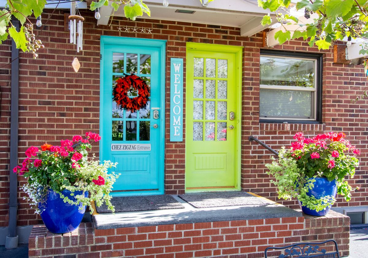 A brick building with a turquoise and lime green door, a 'WELCOME' sign, and a 'Chez ZIGZAG Entrance' sign.