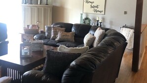 Una sala acogedora con un sofá de cuero oscuro, una mesa de centro de madera y un armario blanco. Un letrero en la pared dice 'Home Sweet Home'.
