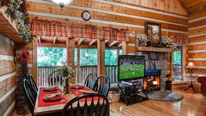 A cozy cabin interior with a dining area, a fireplace, and a television displaying a sports game. - MOUNTAIN HIDEAWAY - AUTHENTIC LOG CABIN MINUTES TO PARKWAY AND ATTRACTIONS!!! (Pigeon Forge)