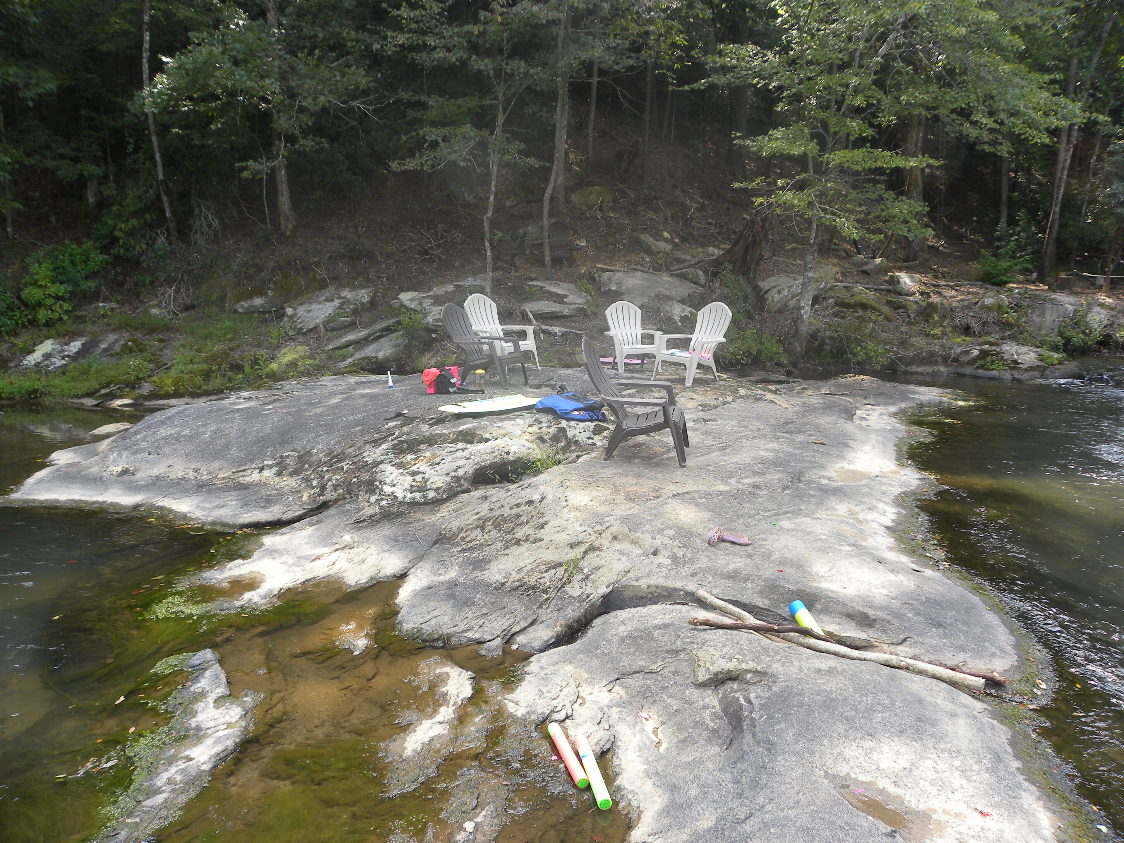 Un cadre naturel avec une formation rocheuse au bord d’une rivière, entourée d’arbres. On y trouve des chaises Adirondack et du matériel de camping sur les rochers.