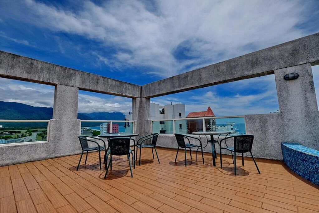 Una terraza en la azotea con suelo de madera, sillas y mesas de metal, que ofrece vistas a un paisaje urbano y montañas.