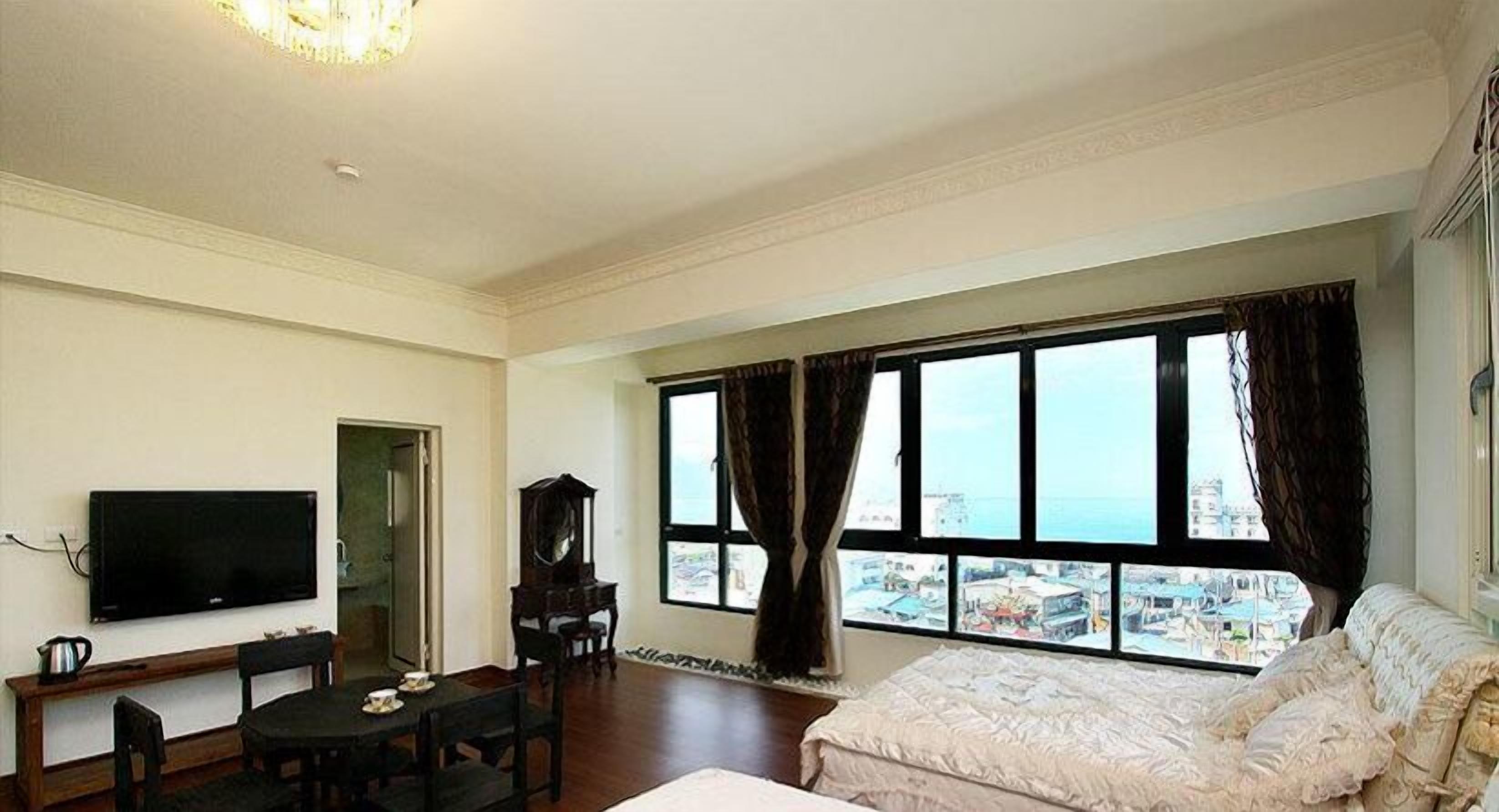 A room with a bed, a TV, a desk, and a view of a cityscape through large windows.