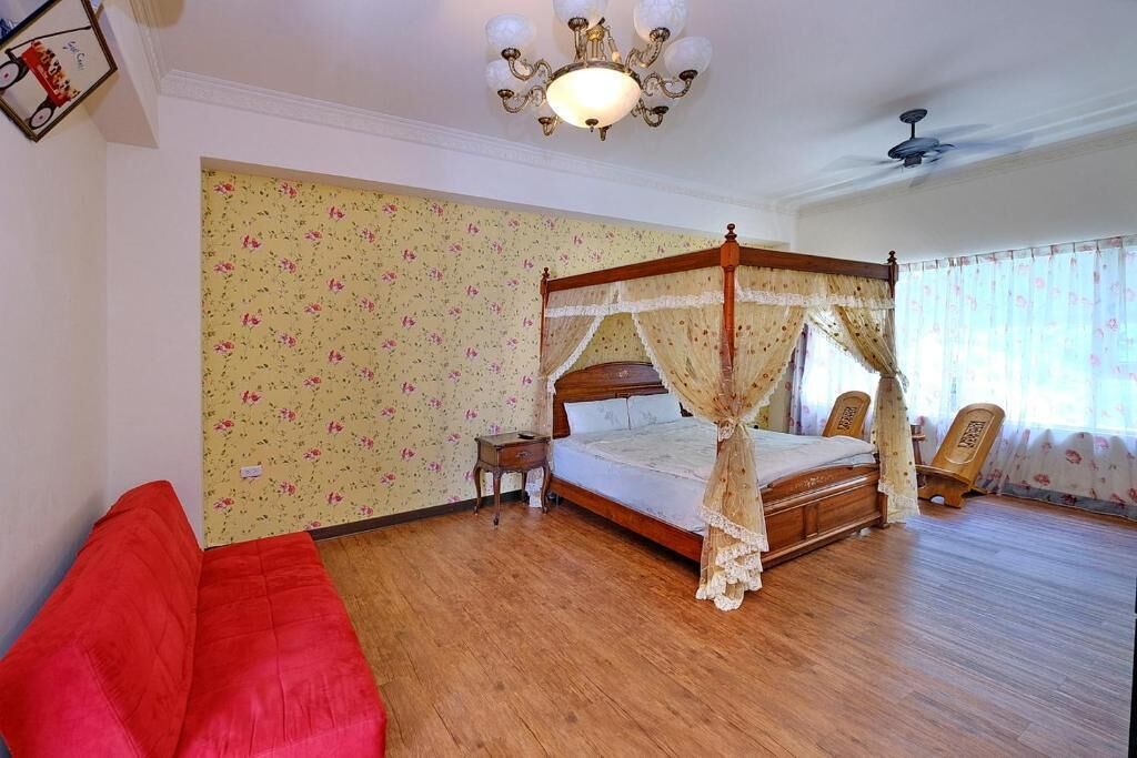 A room with a four-poster bed, a red sofa, a small bedside table, and a ceiling fan.
