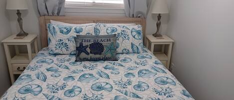 A bed with a blue and white beach-themed bedspread, two bedside tables with lamps, and a pillow with the text "THE BEACH is my happy place."